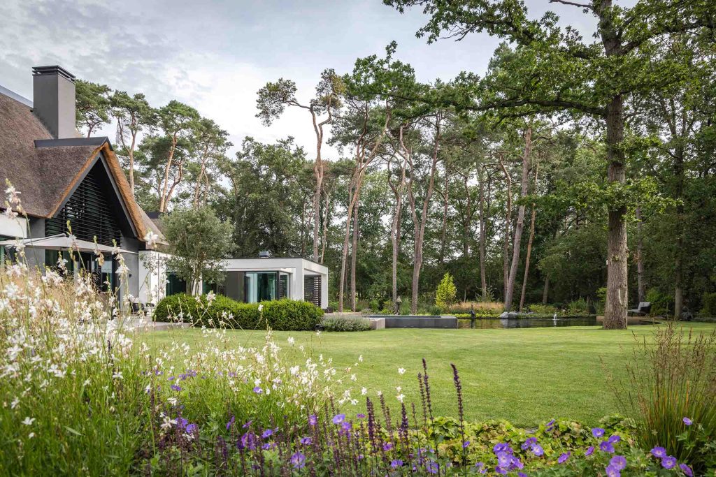 Bosvilla tuinontwerp omgeven door bos met bloeiende planten op de voorgrond. Groot gazon met een fraaie waterpartij voor de villa met rieten dak.