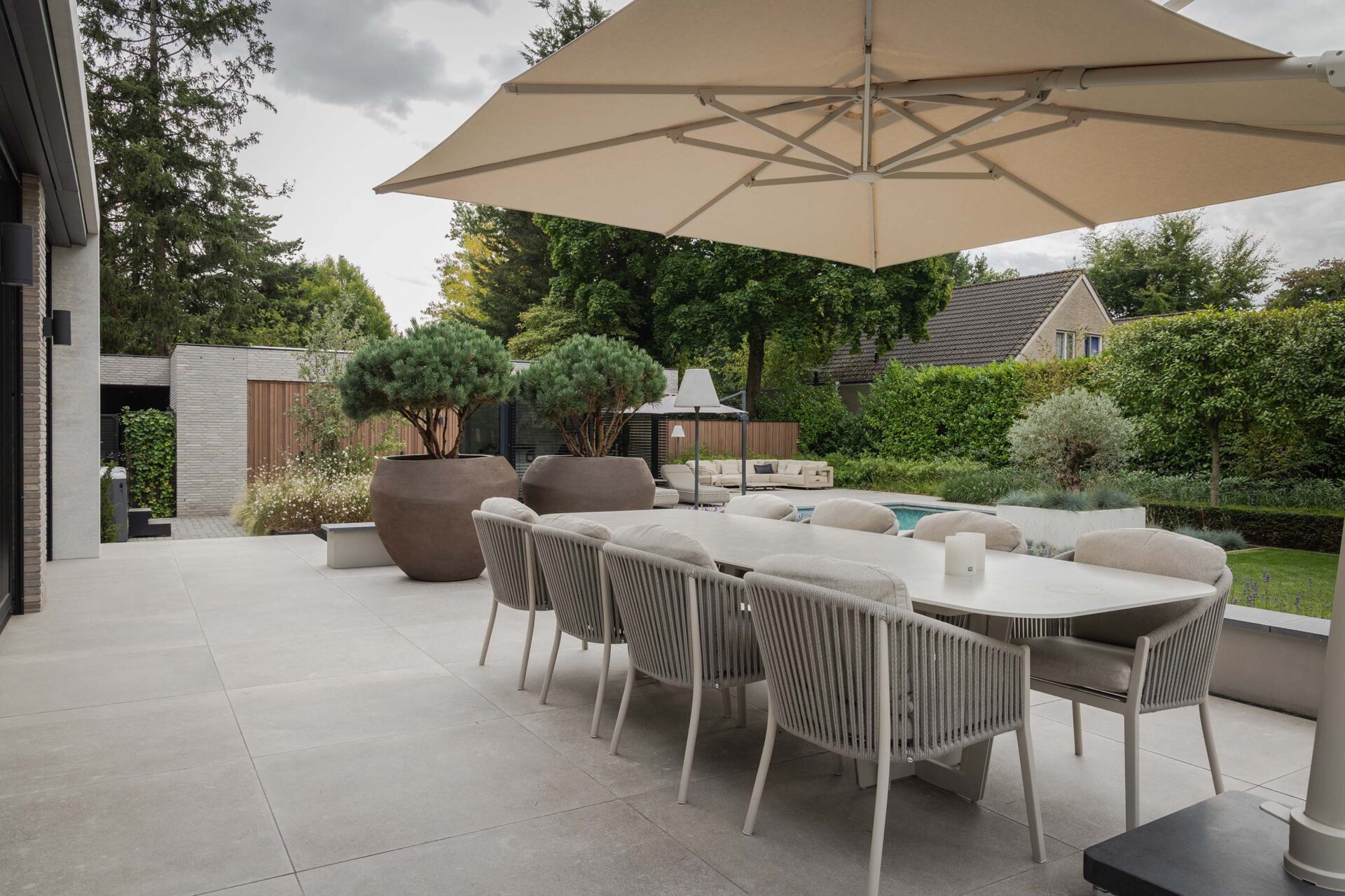 Vollatuin Rosmalen met zicht op het grote terras met grijze grote tegels, lange dinertafel en zeer grote plantenpotten en parasol.