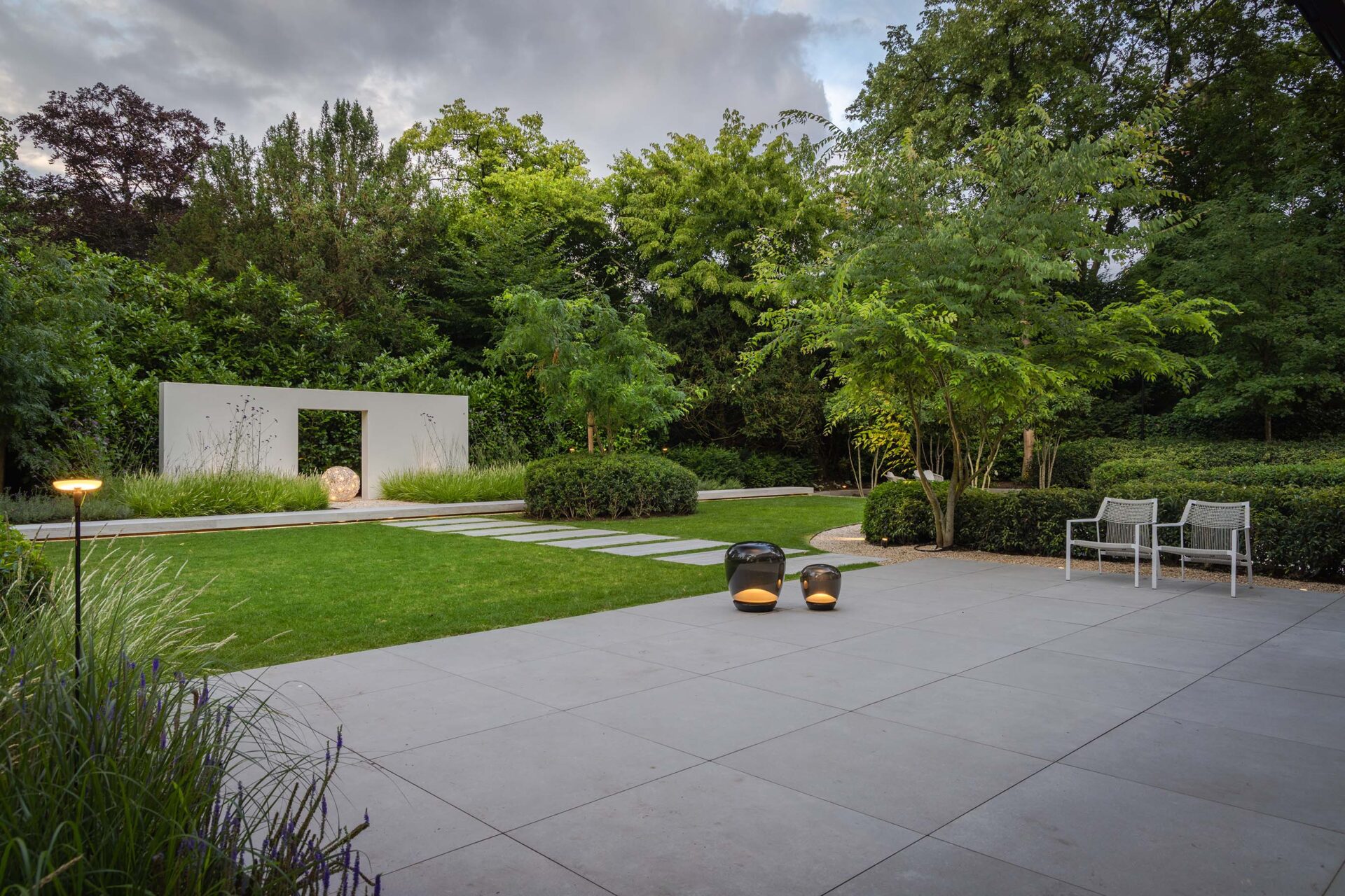 Villatuin Hilversum met zicht op terras met grote grijze tegels en gazon met daarachter een los wit muurelement. 
