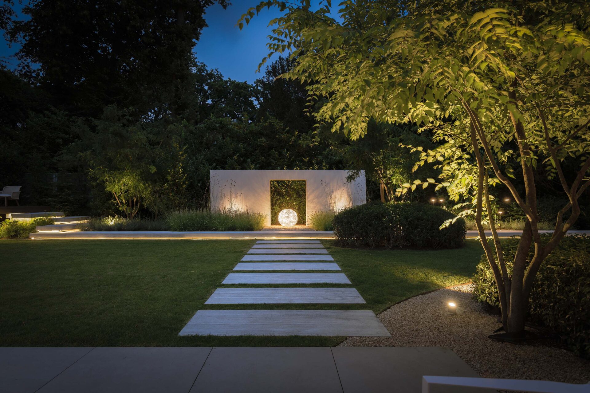Villatuin Hilversum met zicht op rechthoekige stapstenen in het gazon en zicht op een losse witte muur met een opening in het midden en groot rond lichtelement. 