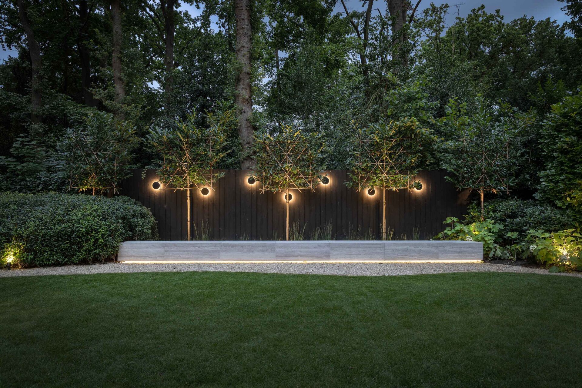 Villatuin Hilversum met zicht over gazon, strakke witte zitrand met verlichting en donkere schutting met ronde designverlichting. 