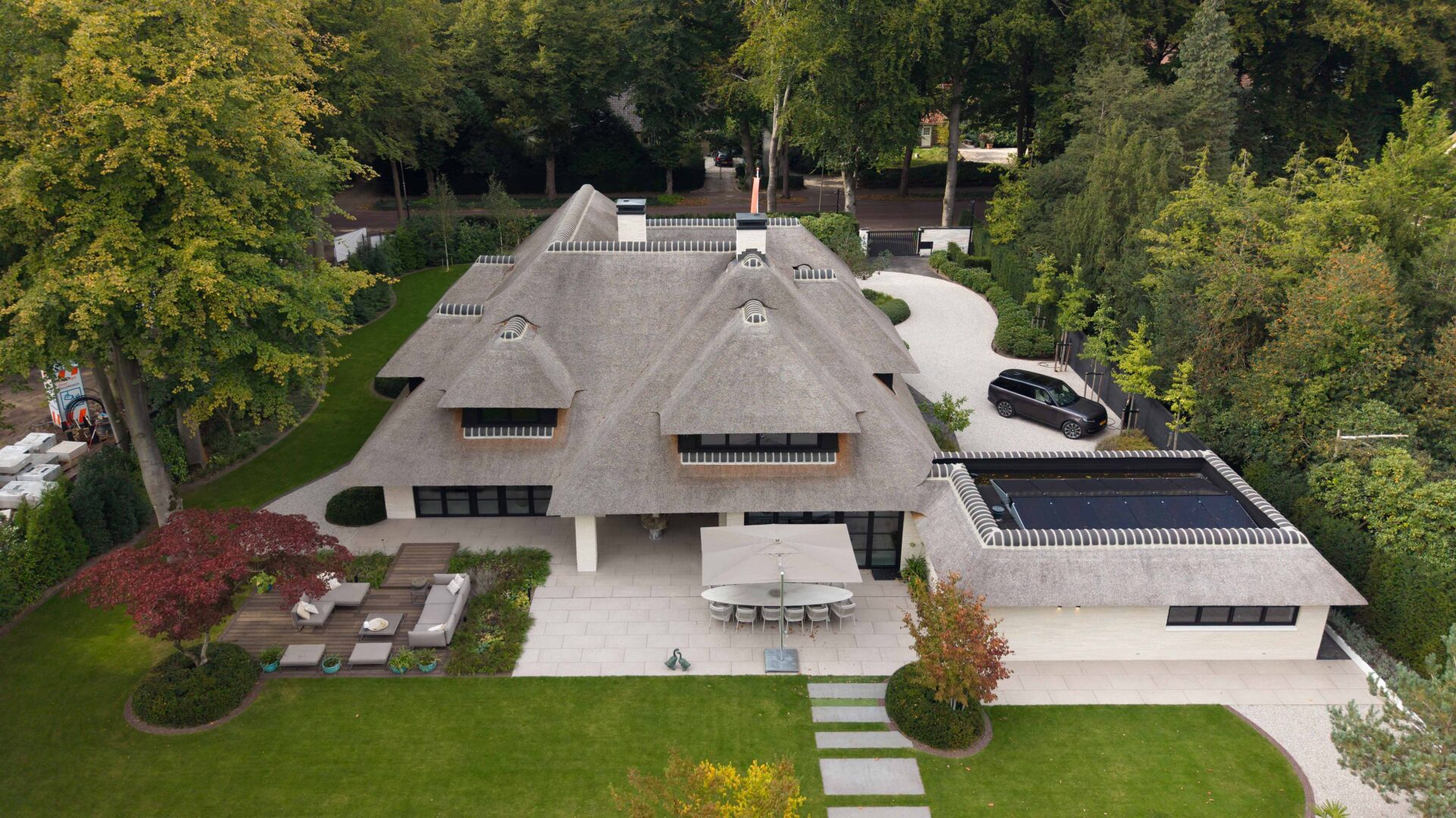 Zicht op de villa te Blaricum in vogelvlucht, rietgedekte villa met groot terras aan de achterzijde met gazon en lichte grote tegels. 